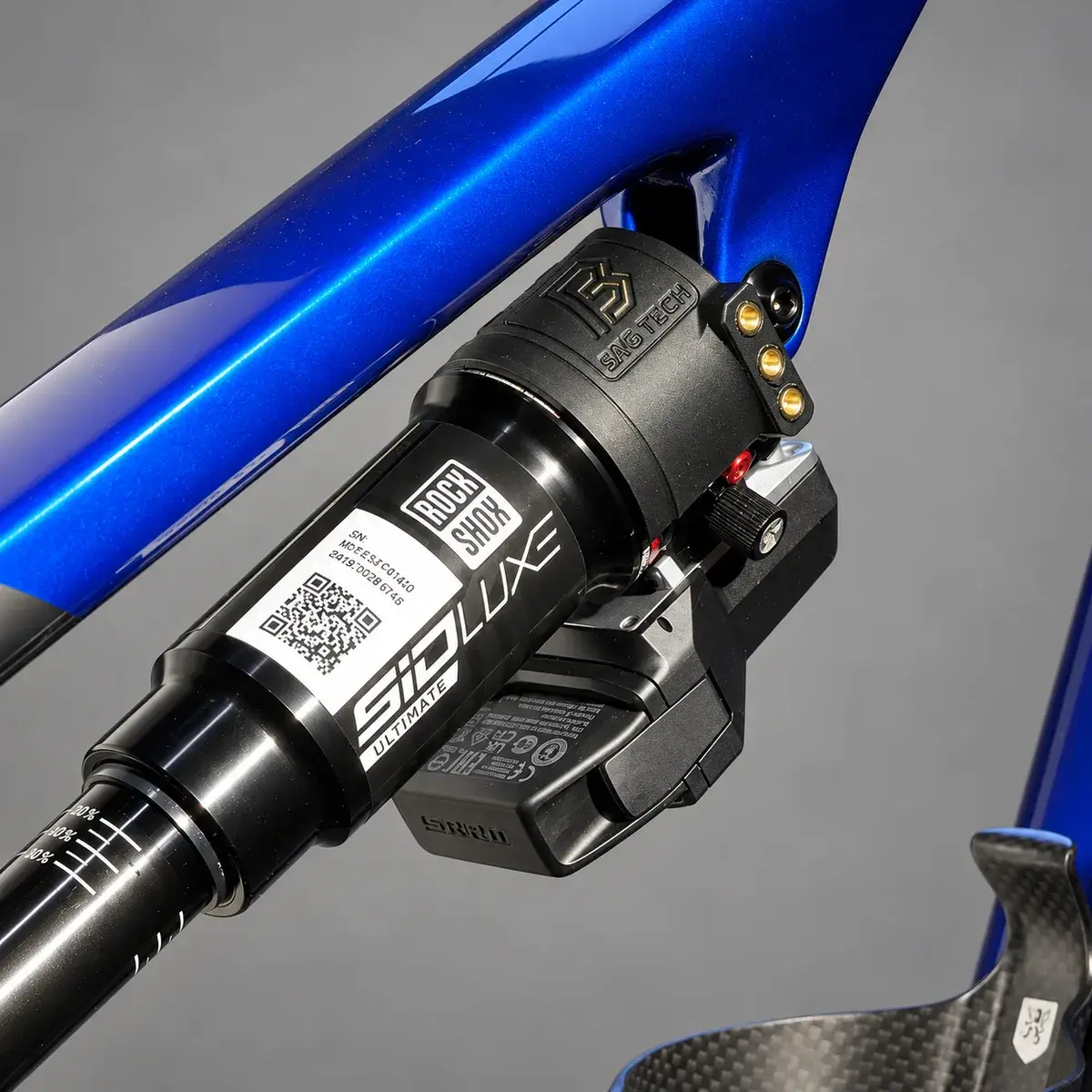A blue frame carrying a RockShox SID Luxe Ultimate shock, with a black BYB Tech sensor mount around the shock body beside the Flight Attendant module, against a grey background.