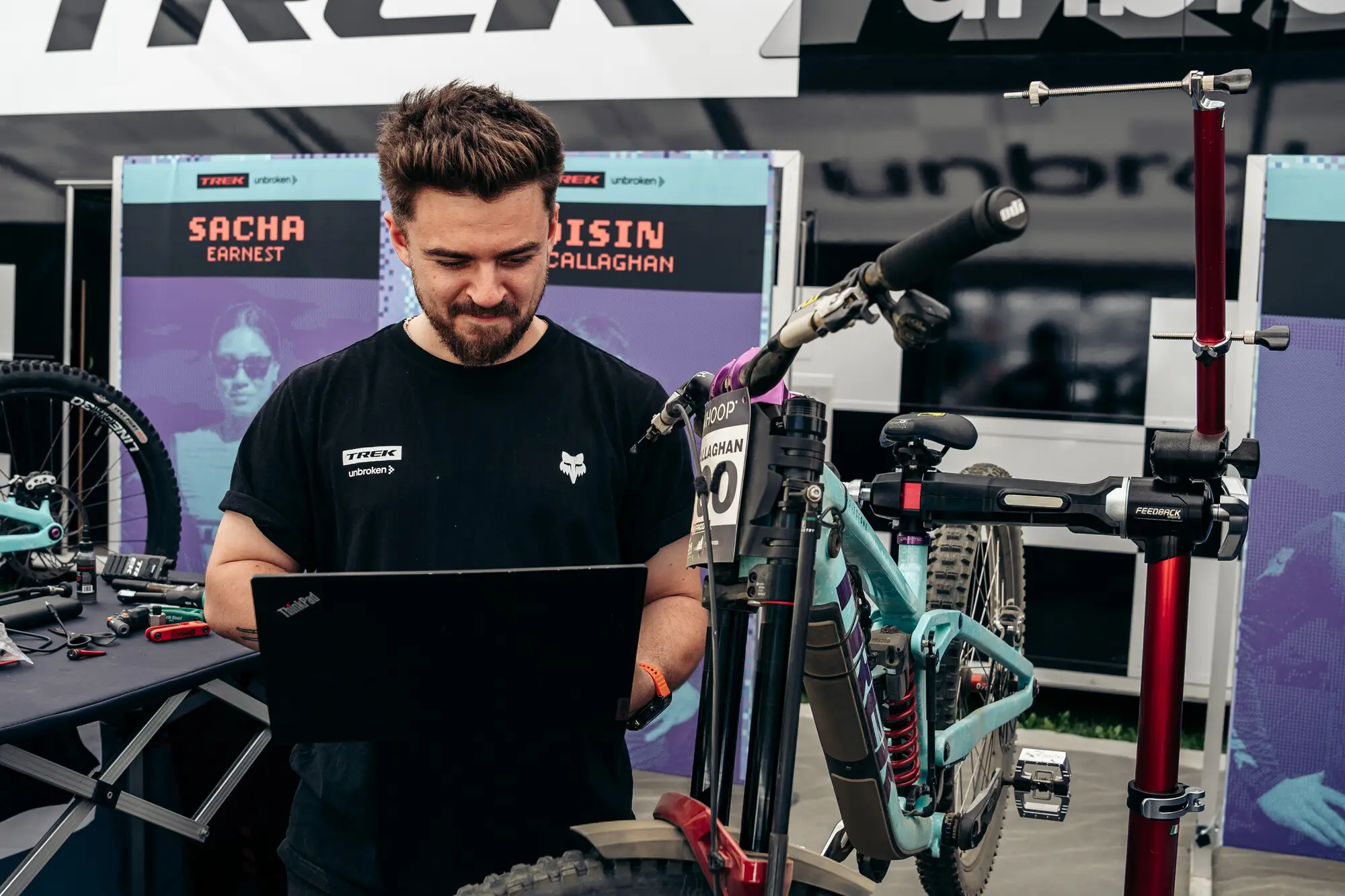 A mechanic reading run data on a laptop in the pits at a UCI Downhill World Cup round, beside a downhill bike in a workstand.