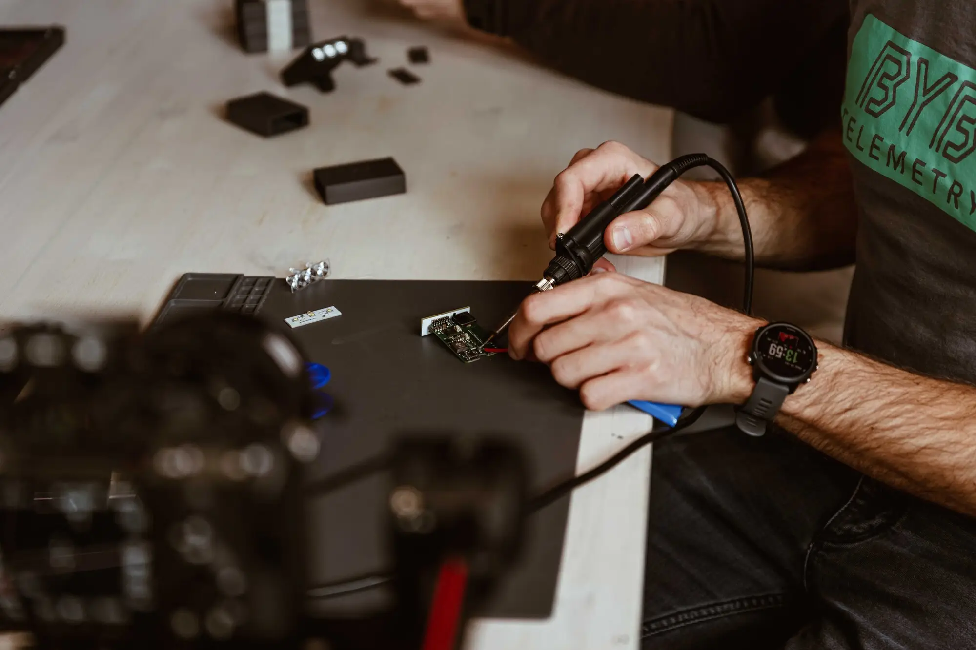 Soldering a component onto a small circuit board at the BYB Tech workbench, with 3D-printed enclosure prototypes and a hot-air tool beside it.