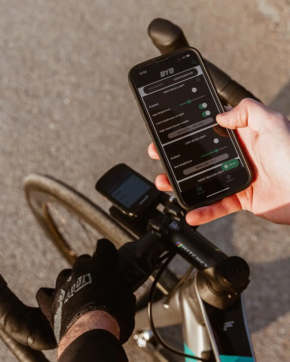 A rider holding a phone above a road bike's handlebar, the BYB app open on the Focus settings with brightness sliders for high and low beam.