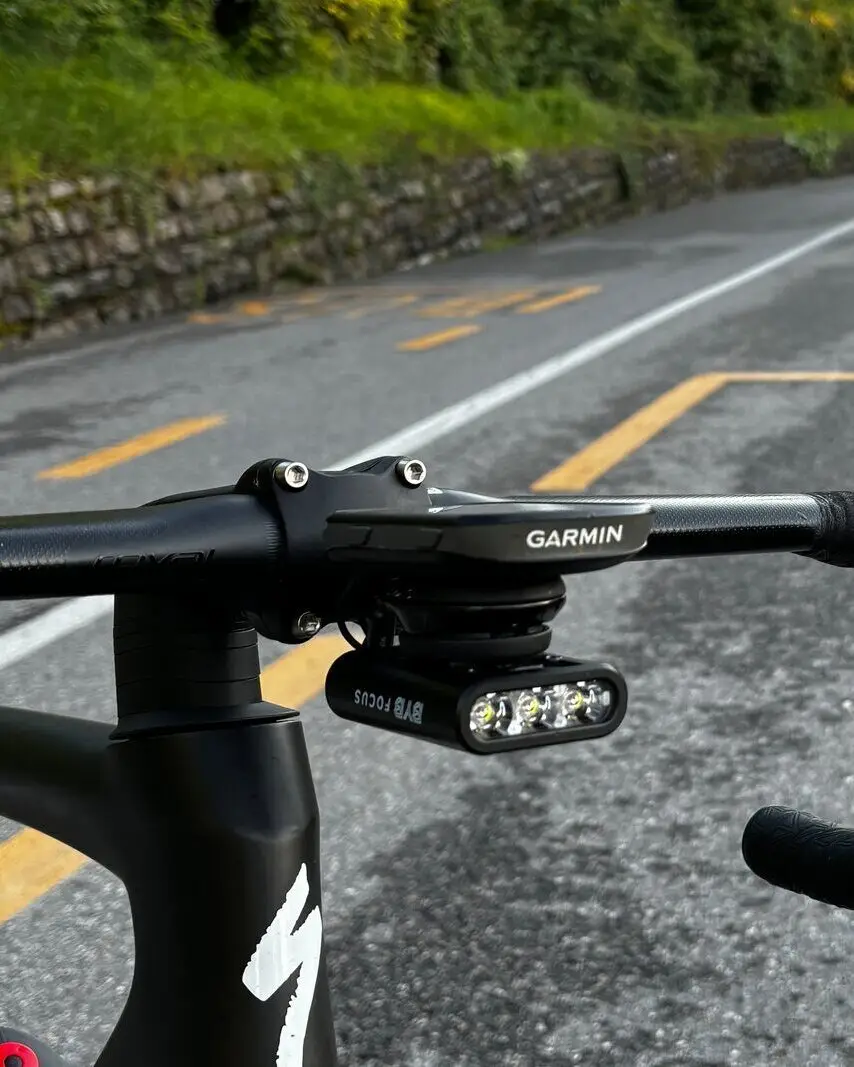 BYB Focus hung under an out-front computer mount on a road bike's handlebar, above a wet country road.
