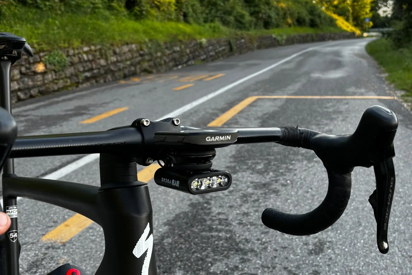 A rider's view over a road bike's handlebar at golden hour, BYB Focus mounted beneath the out-front computer mount, on a wet road.