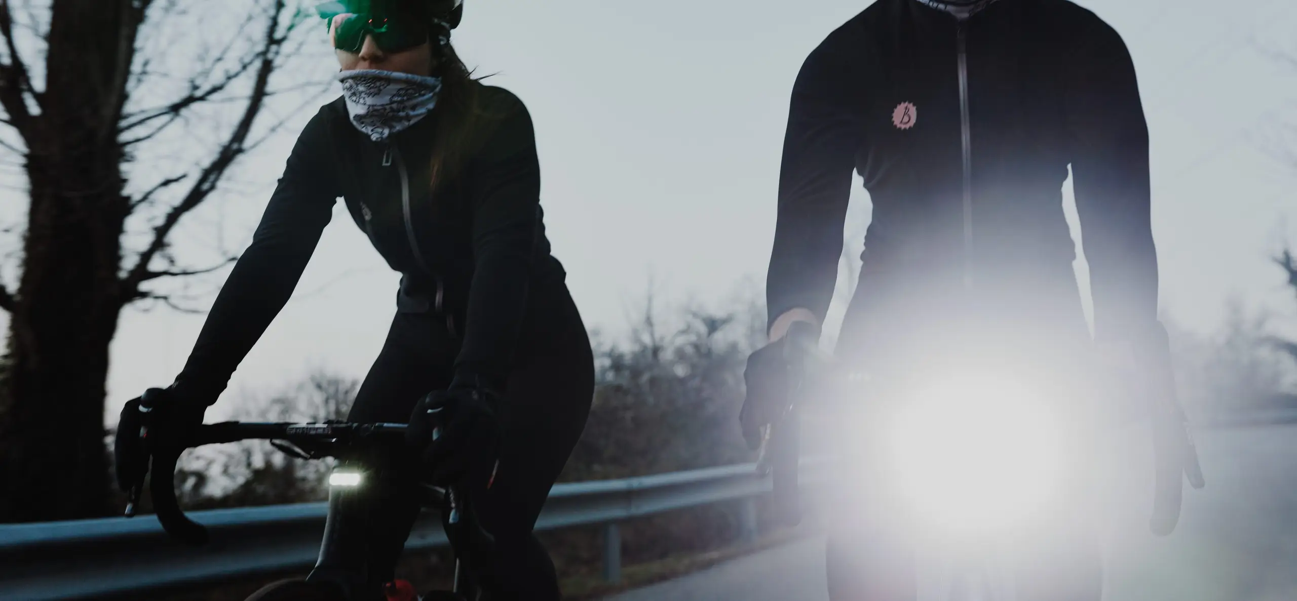 Two road cyclists riding a country road at dusk, the BYB Focus on the right-hand rider's handlebar firing its high beam at the camera.