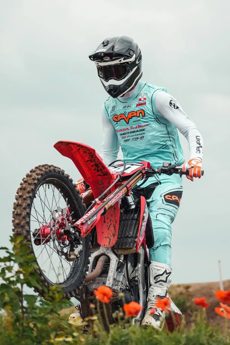 A rider cresting a rise on a red Honda motocross bike, the rear wheel still climbing as the front comes over, with poppies in the foreground.