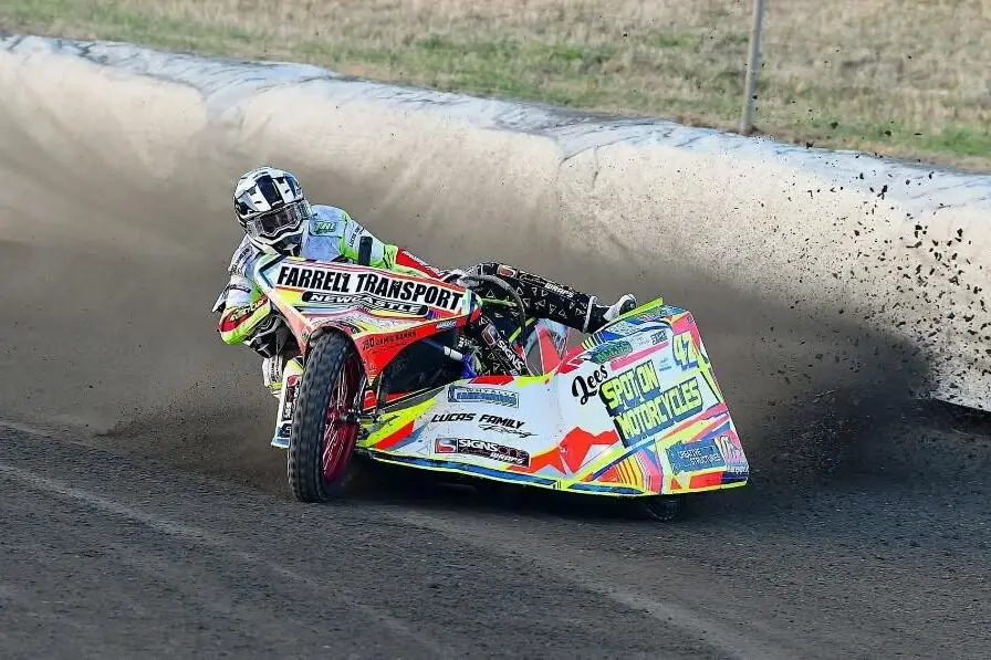 Un sidecar da speedway di traverso in curva su una pista sterrata, con la terra che schizza dalla ruota posteriore.