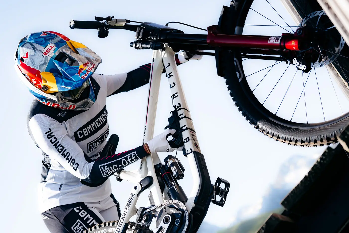 A downhill racer in a Red Bull-branded helmet and white Commencal team kit lifting his bike by the front wheel and top tube, mountains and open sky behind him.