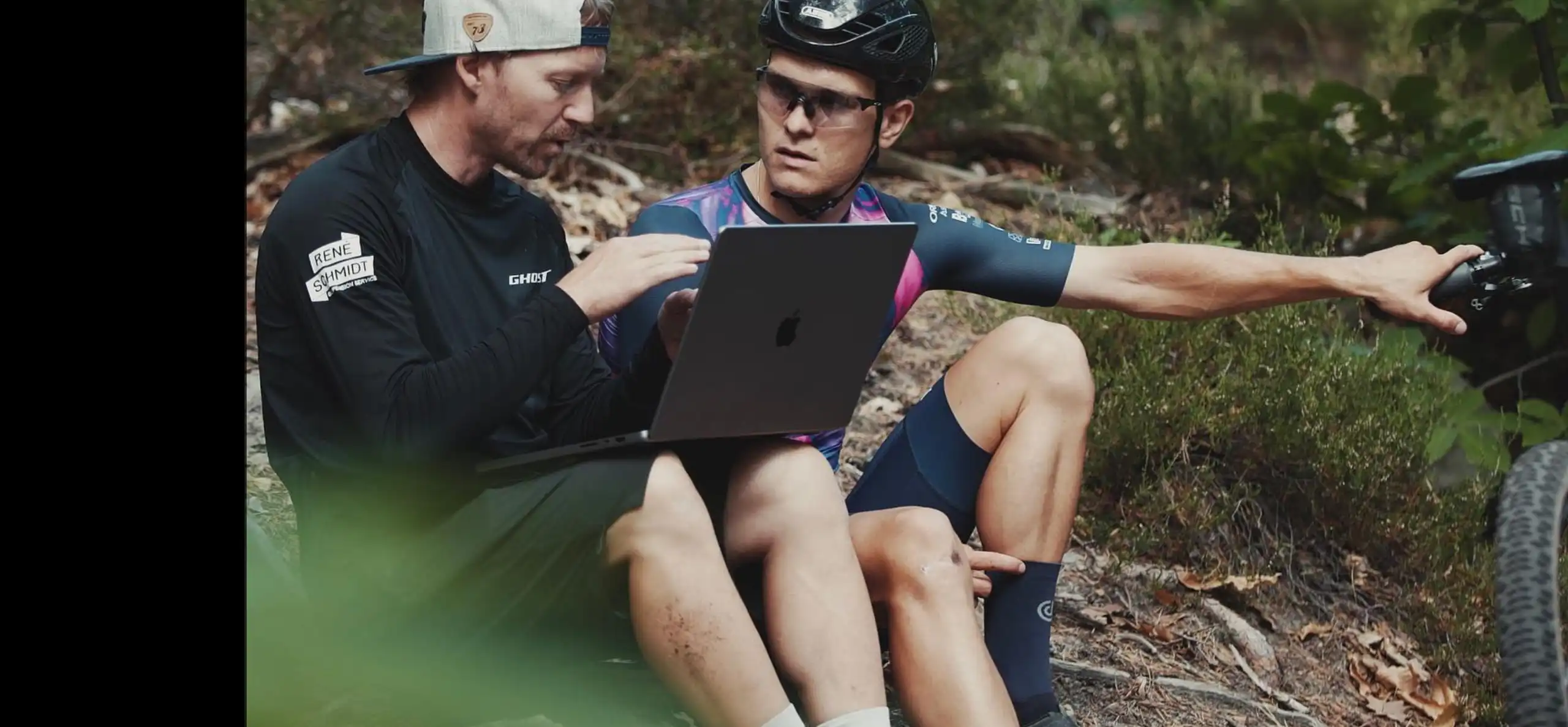 A suspension technician and a mountain bike rider reviewing a recorded session on a laptop at the trailside, the rider pointing back at his bike.