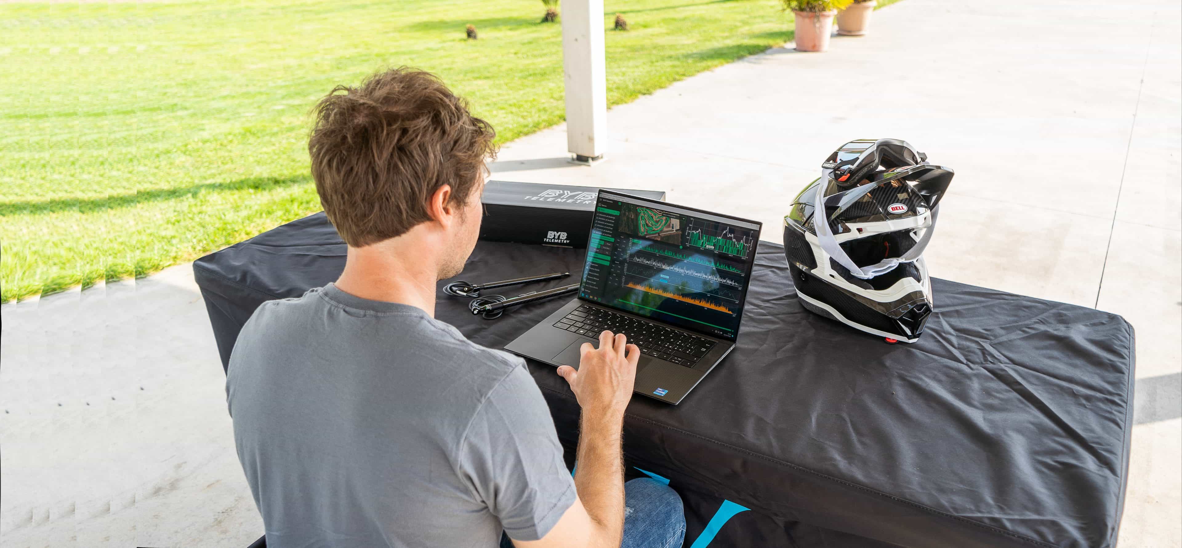 The BYB Telemetry desktop software open on a laptop at a motocross track, suspension traces on screen, beside a helmet and the BYB Telemetry acquisition kit.