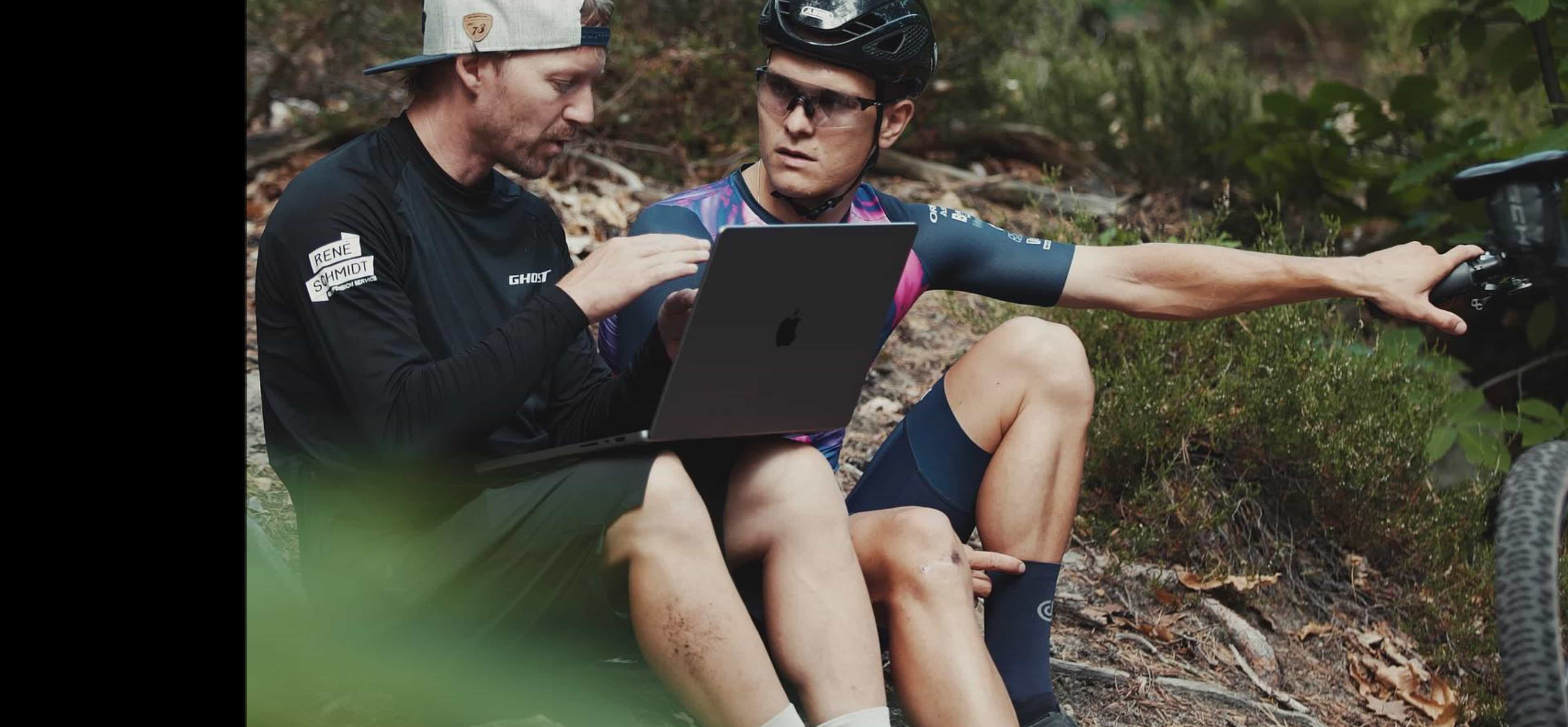 A suspension technician and a mountain bike rider reviewing a recorded session on a laptop at the trailside, the rider pointing back at his bike.