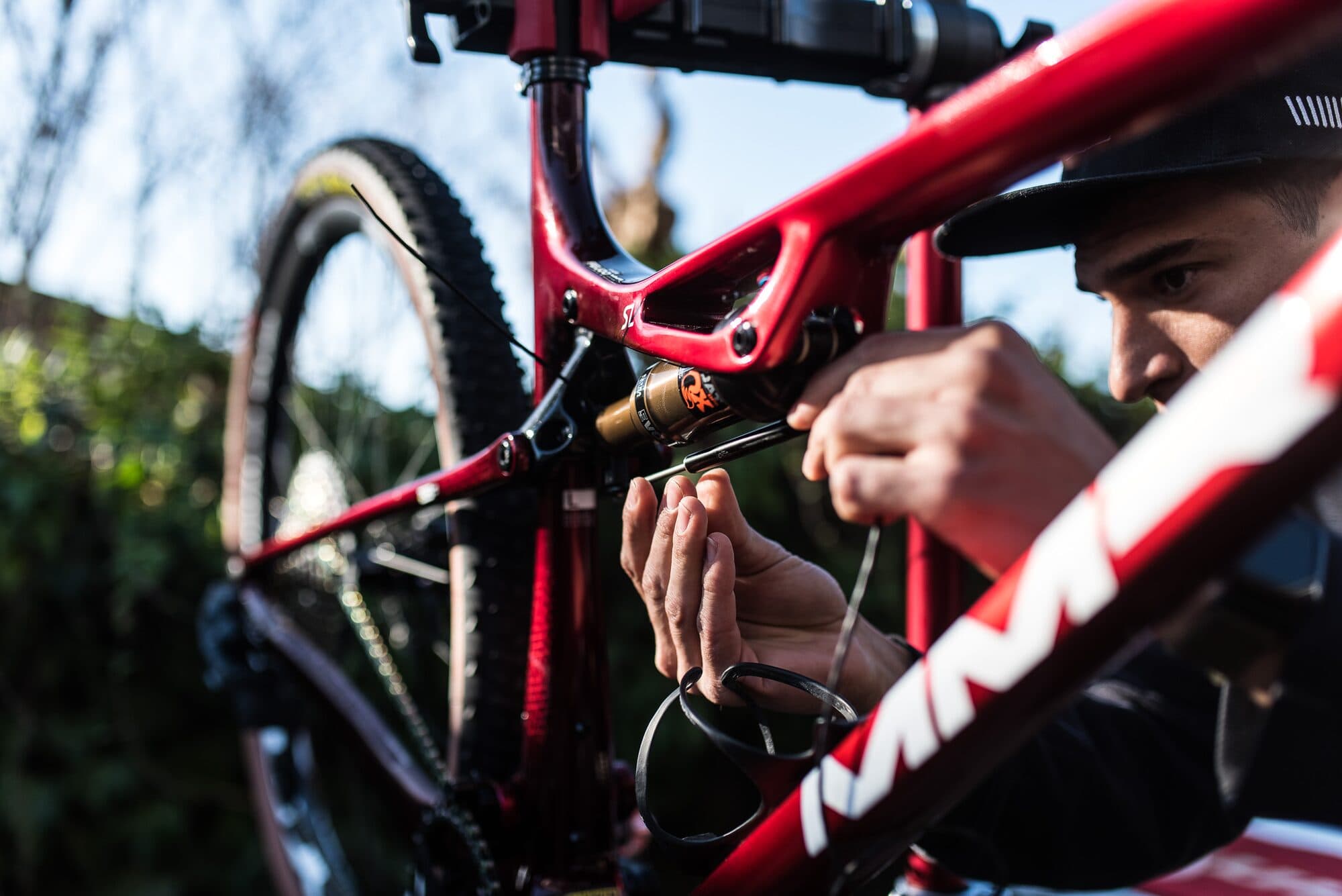 A technician fitting a BYB Tech position sensor alongside the rear shock of a red full-suspension mountain bike, held on a workstand outdoors.