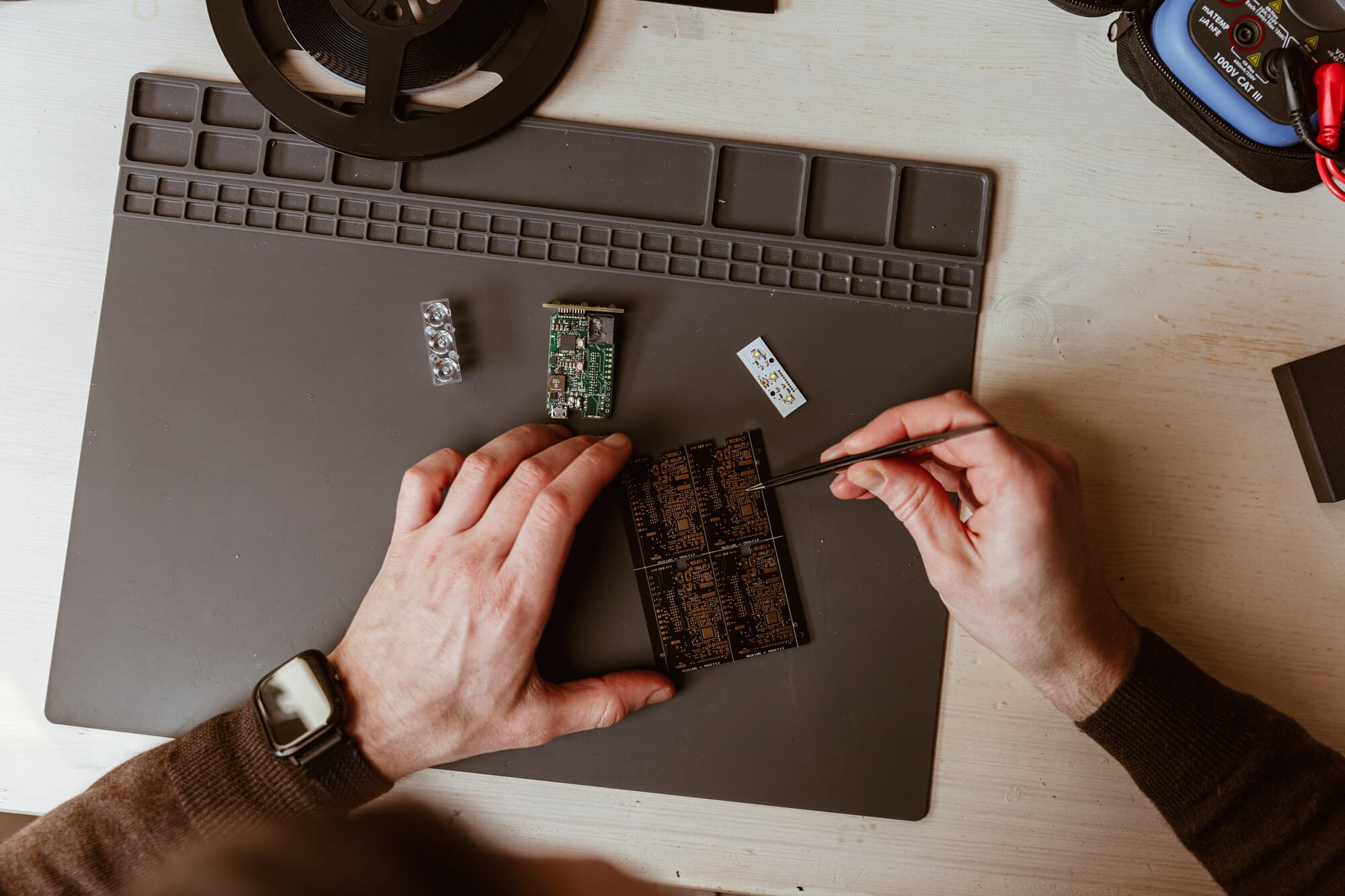 Hands placing a component with tweezers onto a panel of bare sensor boards at the BYB Tech workbench, beside a populated board, an LED board and a reel of parts.