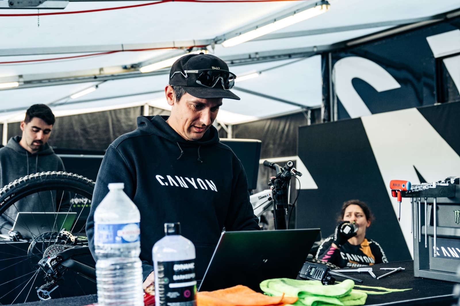 A race team technician reading run data on a laptop in a World Cup pit tent, with bikes and tools around the bench.