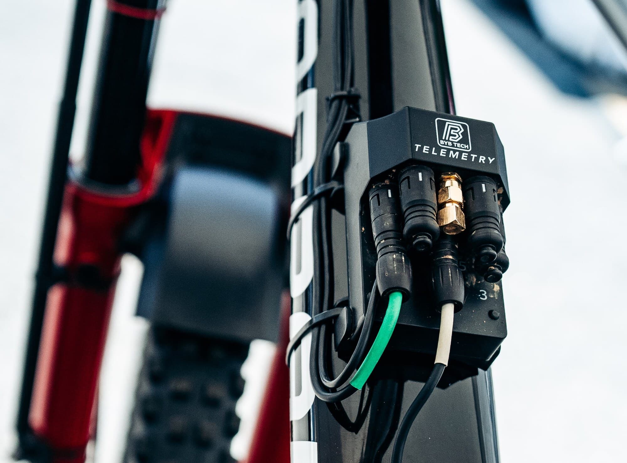 A BYB Telemetry V3 Pro acquisition unit strapped to the frame of a downhill mountain bike, sensor cables plugged into the connectors on its face with a gold GPS antenna fitting among them.