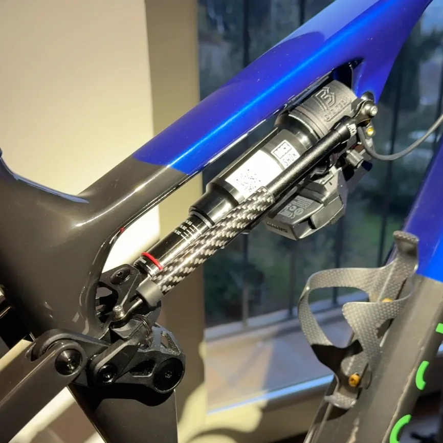 A blue and grey Rockrider Race 940 S indoors by a window, with a linear sensor running diagonally from a printed bracket at the lower linkage.
