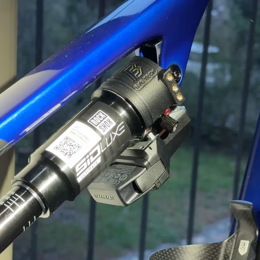 A blue frame carrying a RockShox SID Luxe Ultimate shock, with a black BYB Tech unit clamped around the shock body next to the Flight Attendant module.