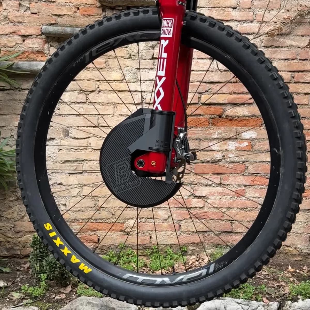 A red RockShox BoXXer fork against a brick wall, with the black printed disc cover fitted over the front rotor and the BYB Tech mark on its face.
