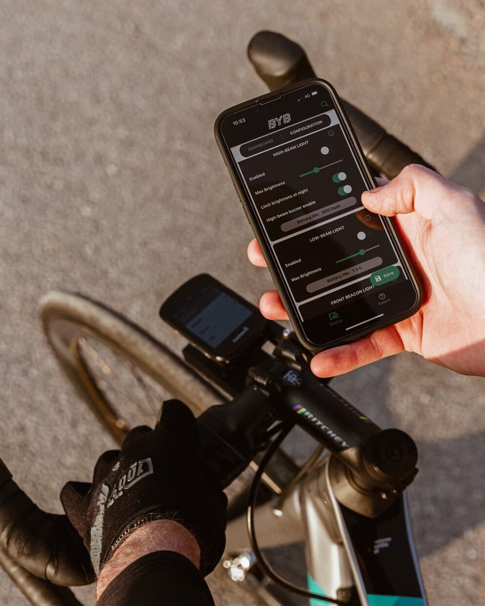 A rider holding a phone above the handlebar with the BYB app open on its configuration screen, showing the high-beam and low-beam sections with brightness sliders, a night-dimming toggle and a battery-life readout.
