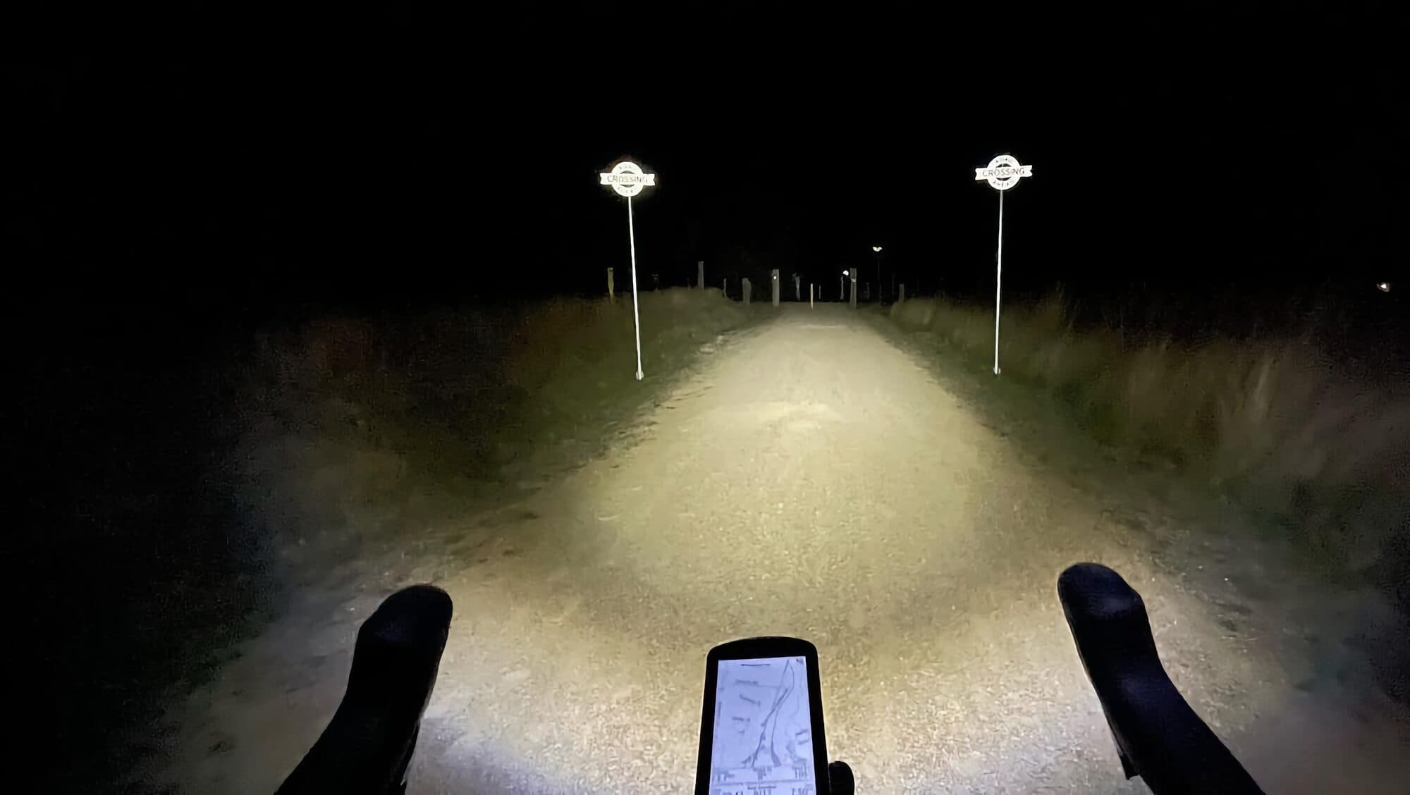 A rider's view over the handlebar at night on an unlit rail trail, the BYB Focus beam lighting the gravel path and two trail signs ahead.