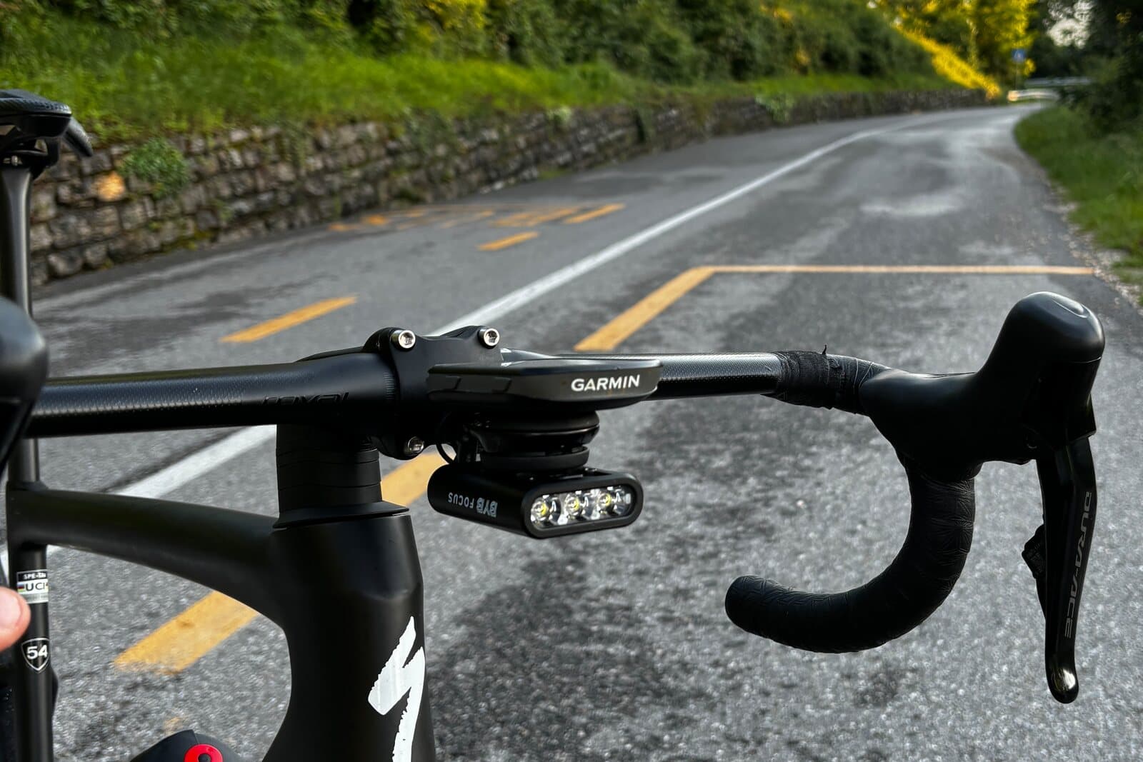 A rider's view over a road bike's handlebar at golden hour, BYB Focus mounted beneath the out-front computer mount, on a wet road.