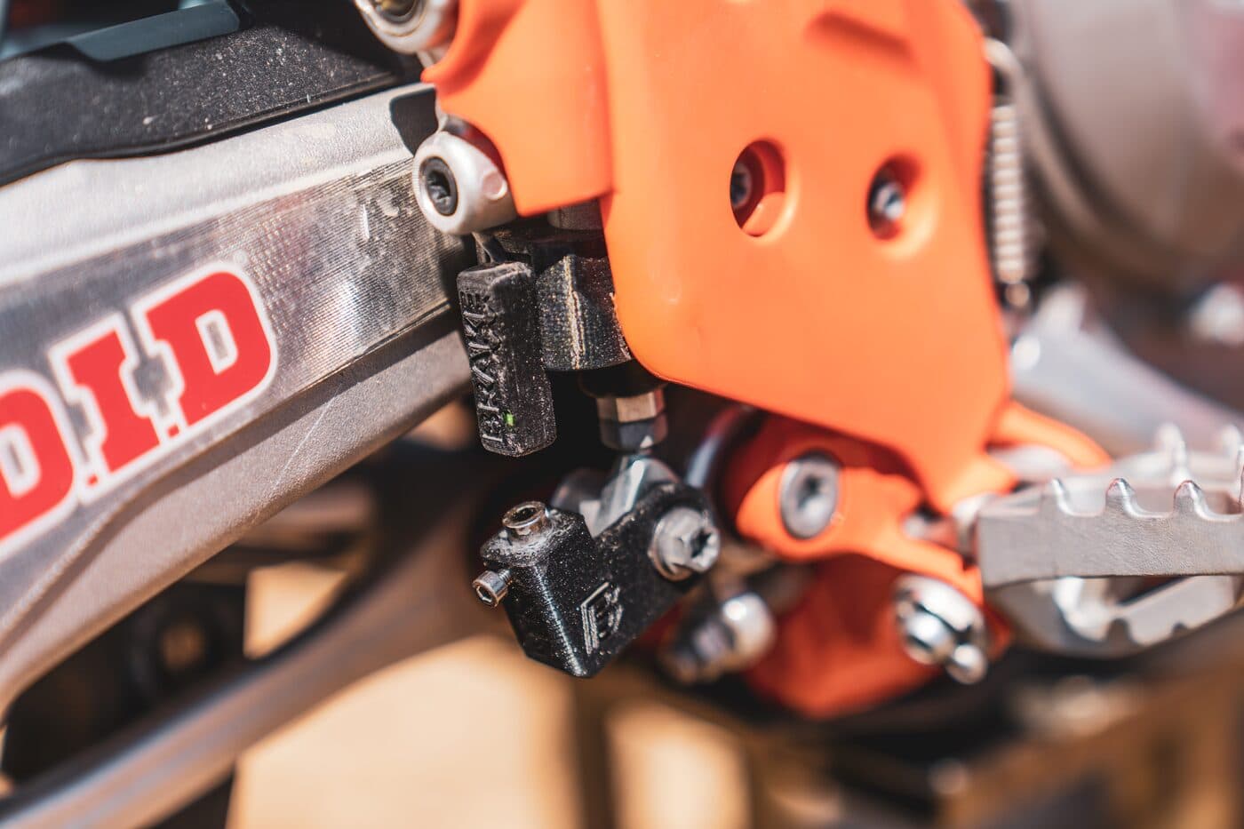 A black BYB Telemetry brake sensor mounted at the rear brake master cylinder of an orange KTM, below the swingarm and beside the footpeg.