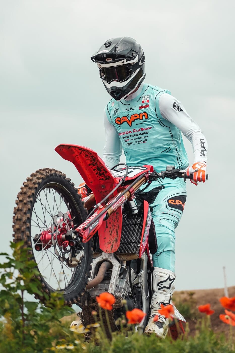 A rider cresting a rise on a red Honda motocross bike, the rear wheel still climbing as the front comes over, with poppies in the foreground.
