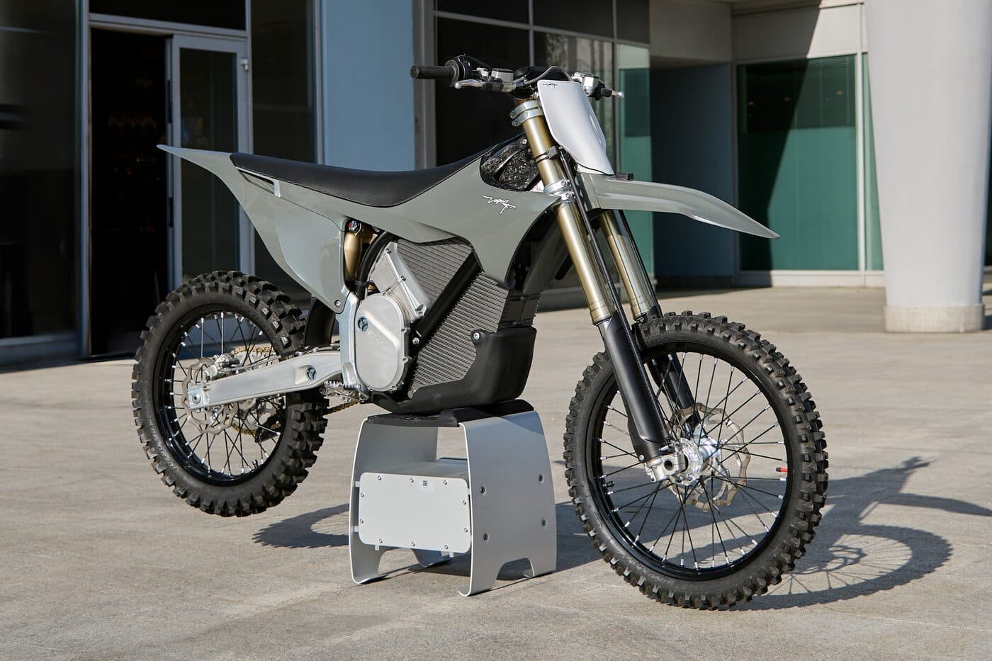 A motocross bike on a paddock stand outside a building, side on, its fork and shock unloaded.