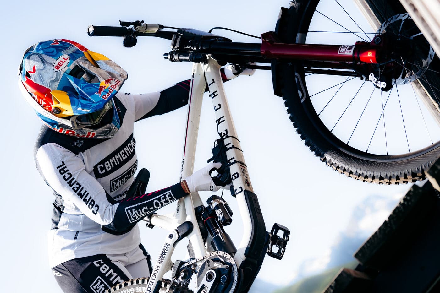 A downhill racer in a Red Bull-branded helmet and white Commencal team kit lifting his bike by the front wheel and top tube, mountains and open sky behind him.