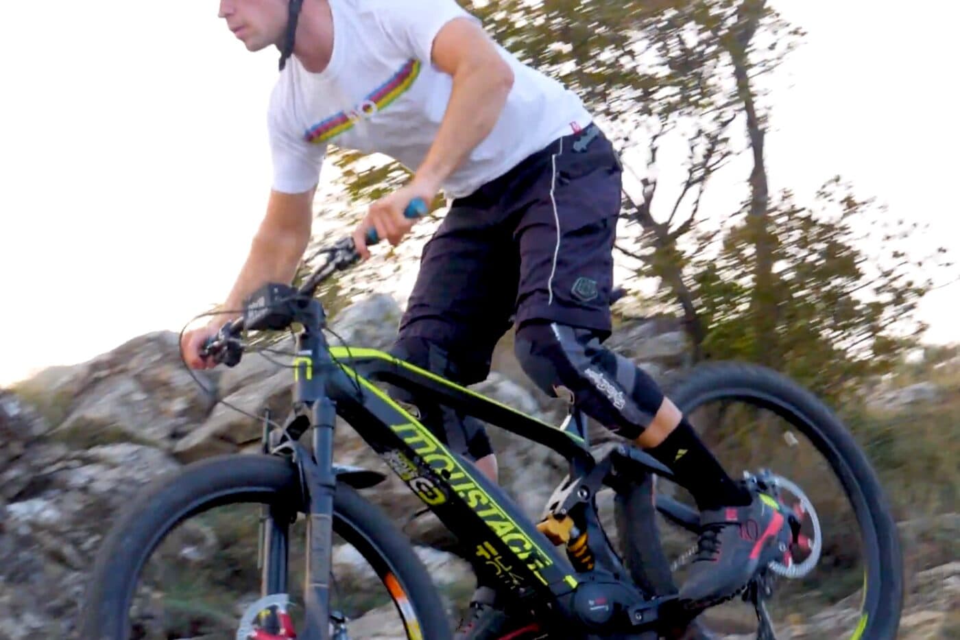 A rider on a Moustache full-suspension e-MTB riding over rock, the motor visible at the cranks and the background blurred with speed.