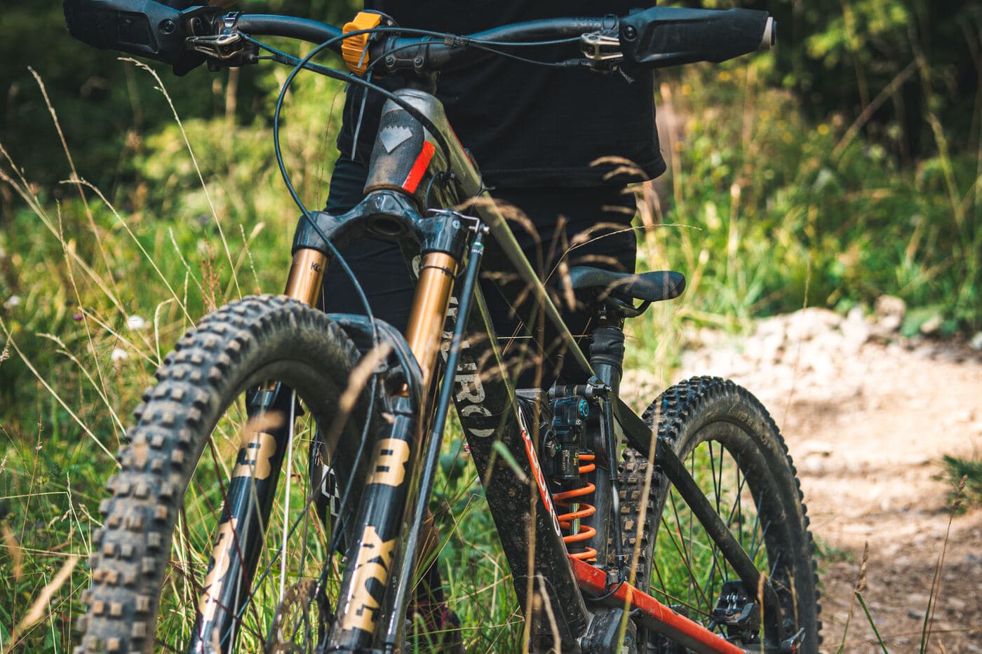 A rider's legs beside a full-suspension mountain bike with gold-anodized fork stanchions and an orange coil shock, standing in tall grass beside a dirt trail.