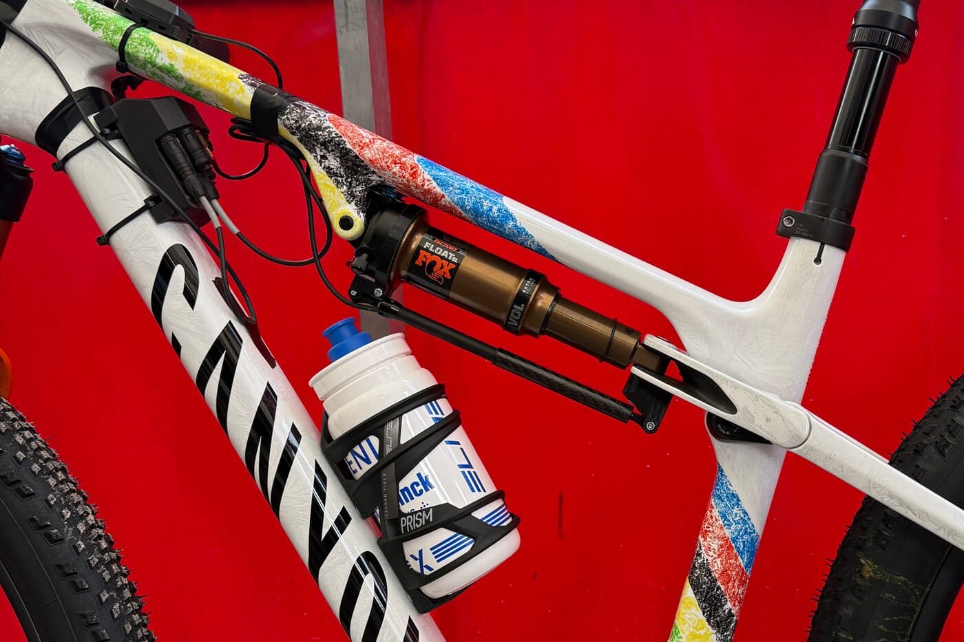 A BYB Telemetry acquisition unit fitted to the top tube of a Canyon cross-country race bike painted in world champion rainbow stripes, with the cabling running down past the Fox shock.