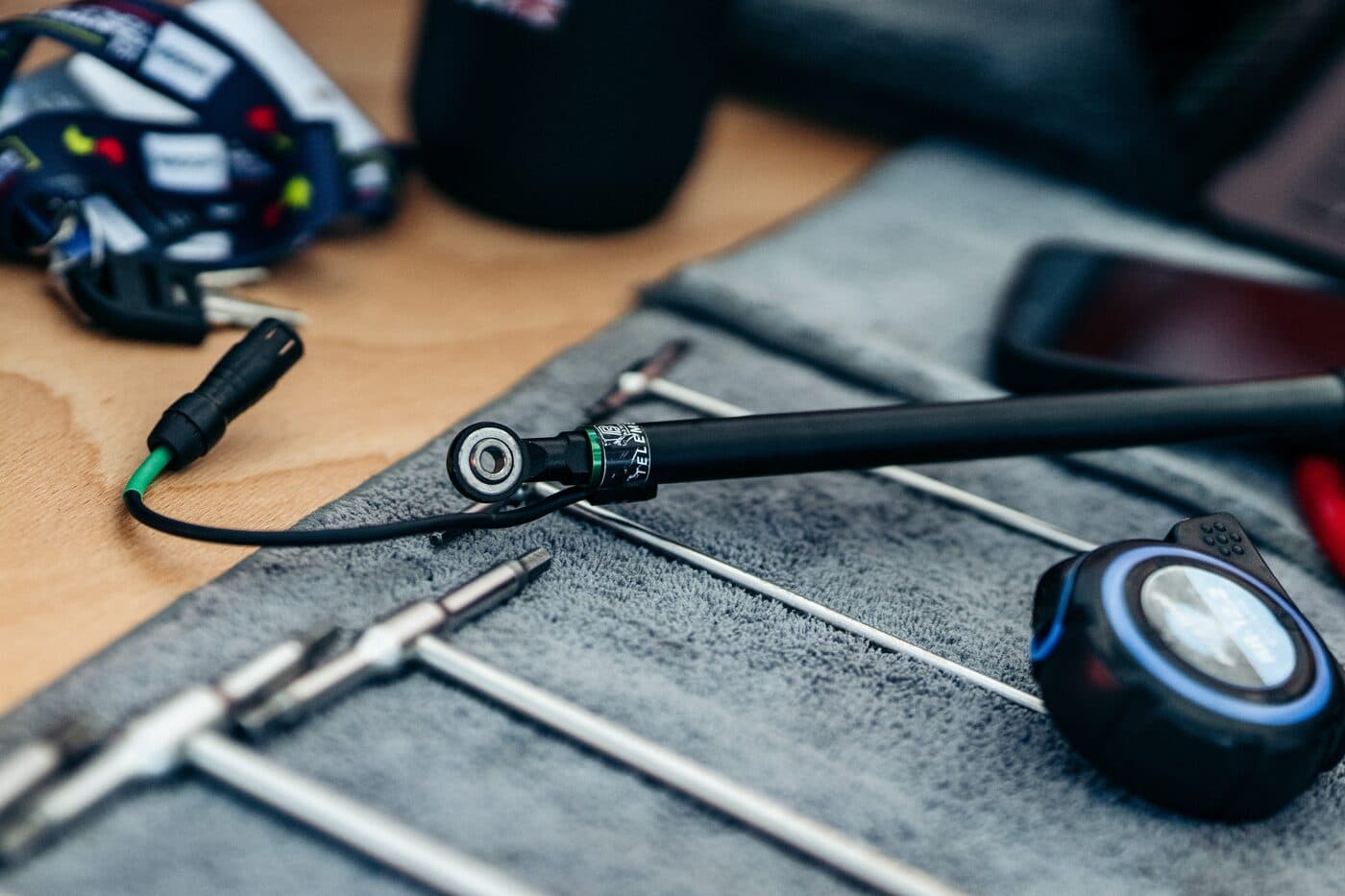 A BYB Telemetry suspension sensor lying off the bike on a mechanic's towel in the pits, its eyelet joint and green-collared cable connector in focus beside a roll of T-handle hex keys.