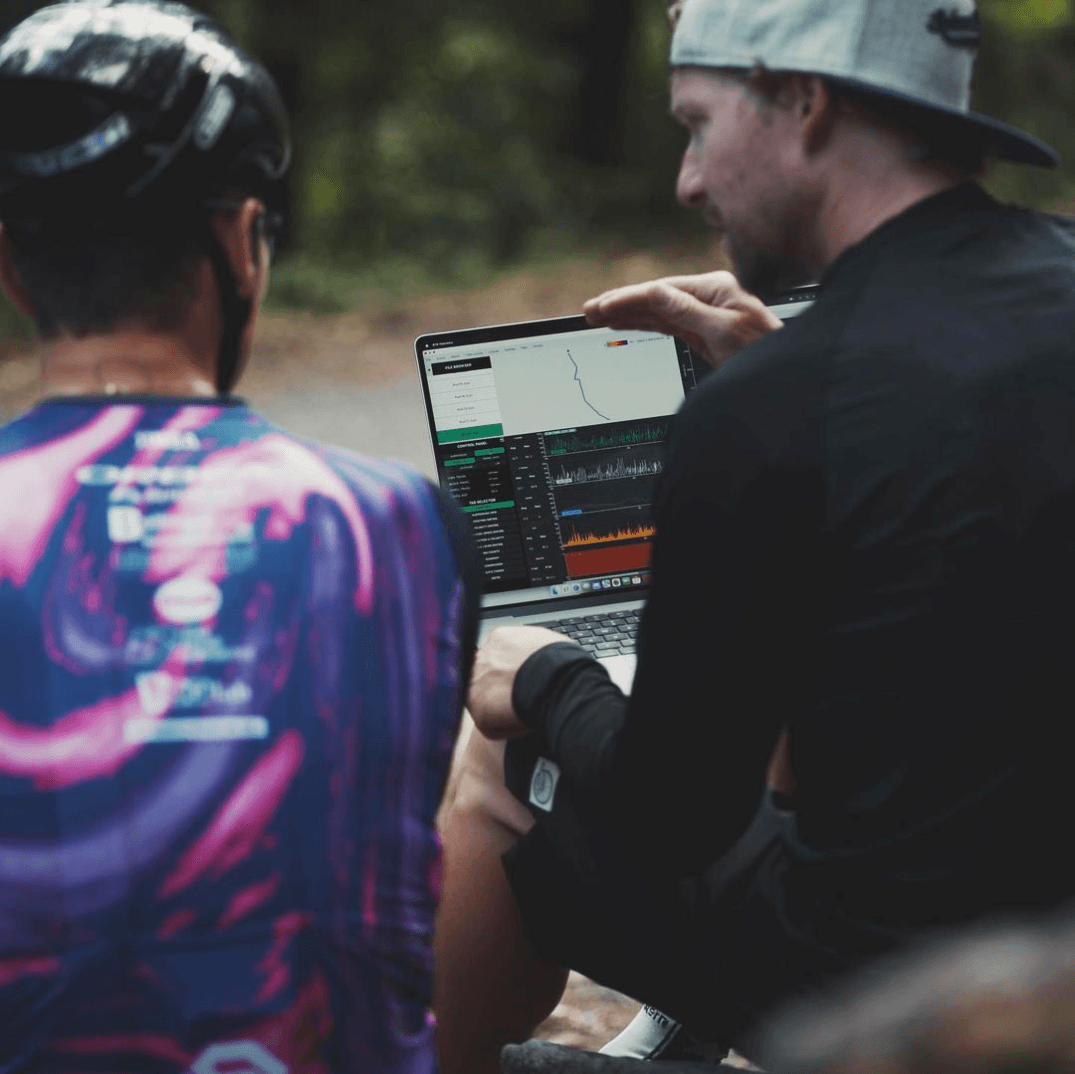 A suspension technician and a rider sitting at the trailside reading a run back on a laptop, the BYB software showing the fork and shock traces from the descent they have just ridden.